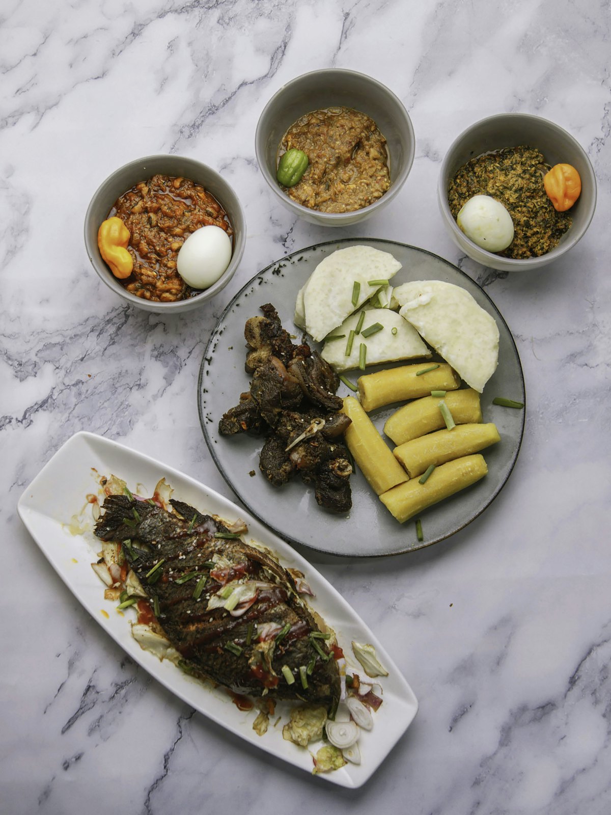 A spread of kenkey, yam and grilled fish with bowls of stew and kontomire